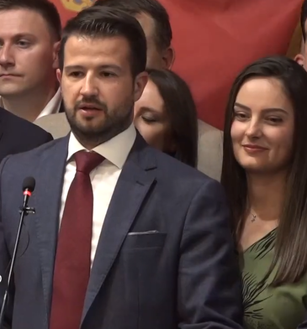 Ein Mann im Anzug und Krawatte steht an einem Rednerpult mit einem Mikrofon und spricht vor einer Menge mit einem Plakat im Hintergrund, wahrscheinlich auf ein Regierungshandeln reagierend.