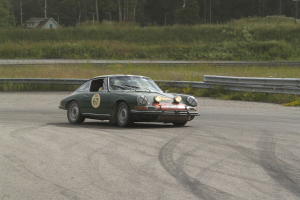Ein grünes Auto fährt auf einer Rennstrecke, umgeben von einem Metallzaun, Pflanzen, Gras, Bäumen, einem Haus mit Dach, Pfählen und einem bewölkten Himmel.