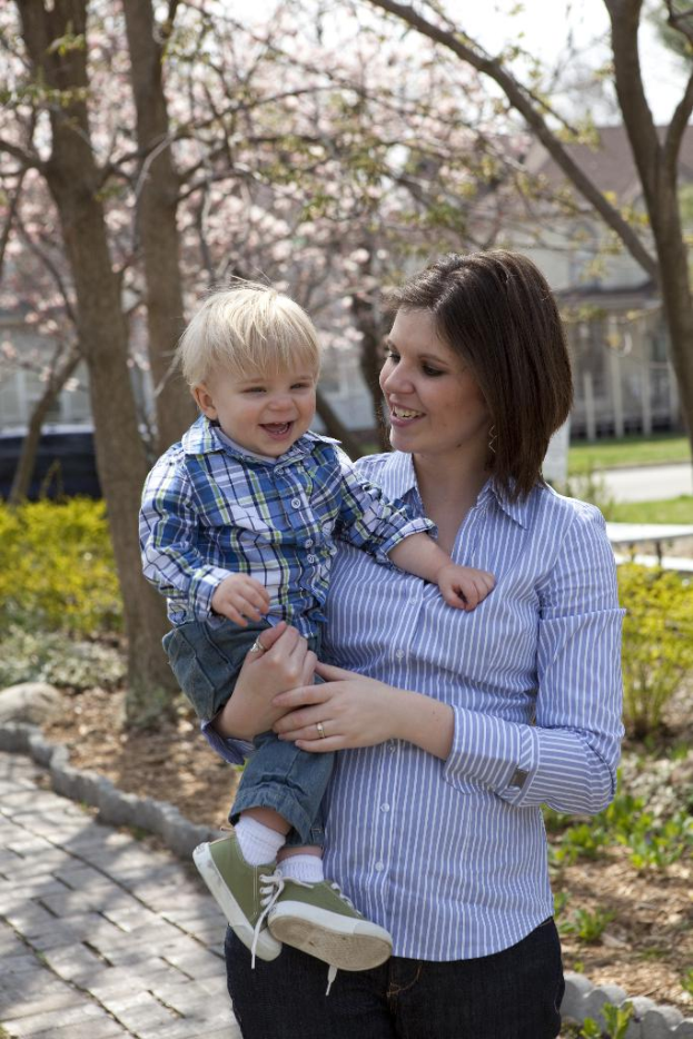 Eine Frau mit einem strahlenden Baby auf dem Arm, beide strahlend, mit Bäumen, Pflanzen, einem Weg, Häusern und Himmel im Hintergrund.